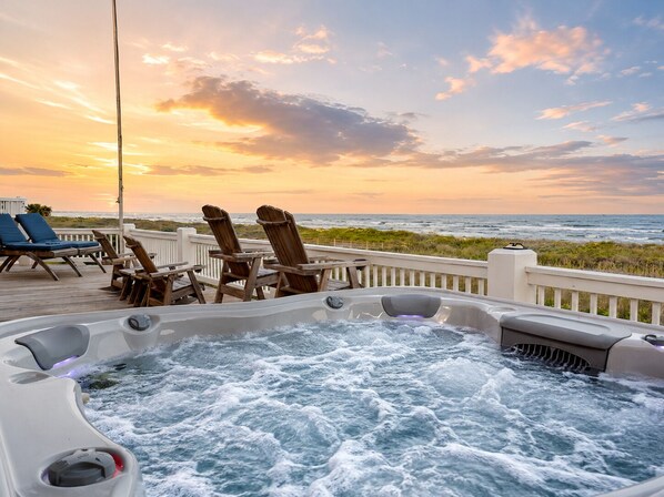 A hot tub with a view of the ocean at sunset, wooden deck chairs, and a railing.