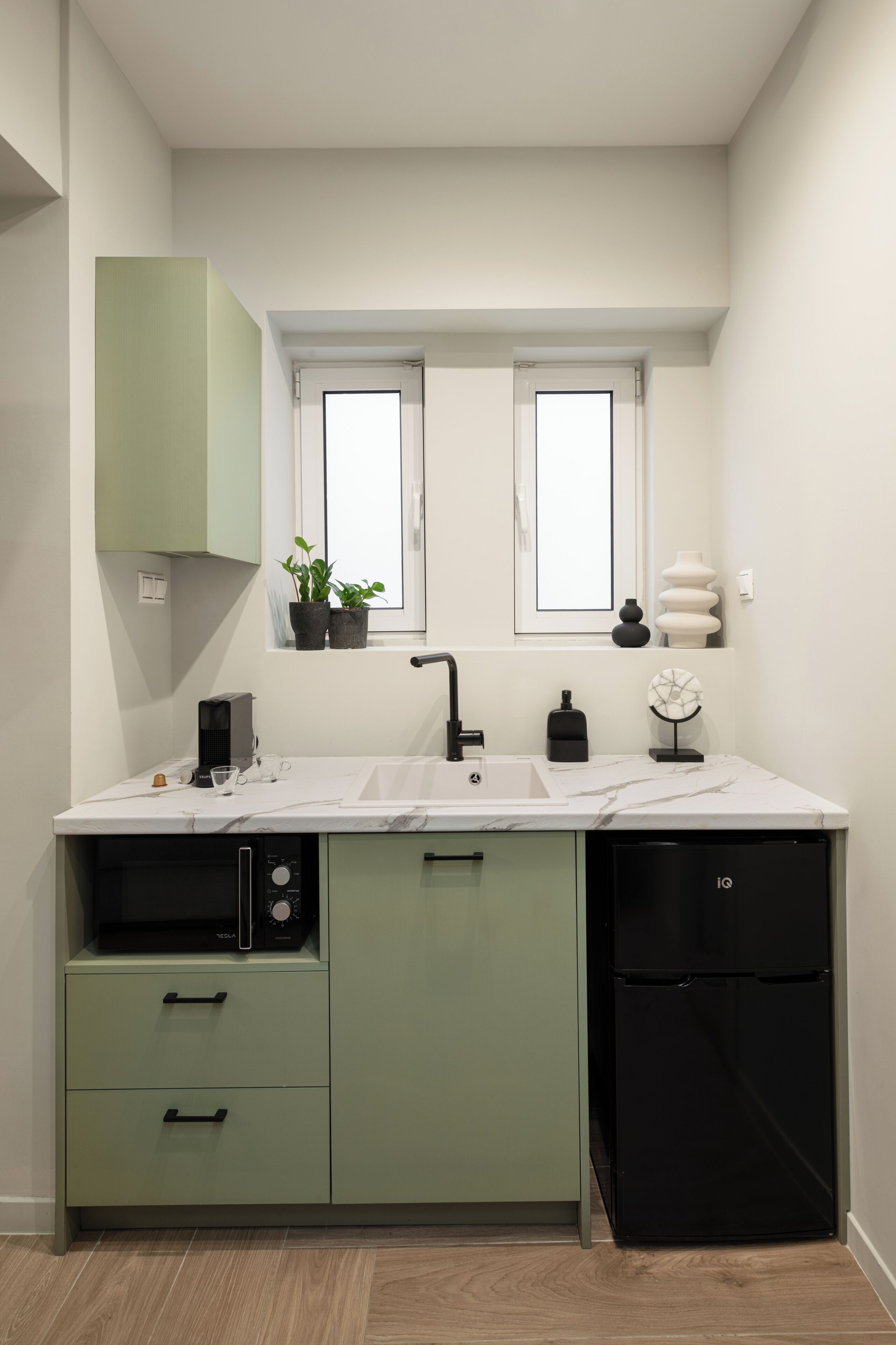 A compact kitchen with a built-in dishwasher, a microwave, and a black refrigerator. There are two windows above the sink, and a small potted plant on the windowsill.