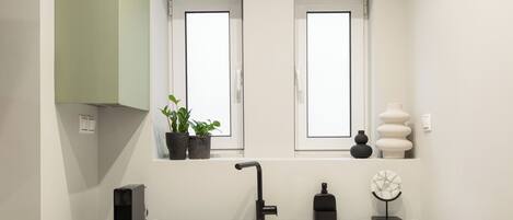 A compact kitchen with a built-in dishwasher, a microwave, and a black refrigerator. There are two windows above the sink, and a small potted plant on the windowsill.