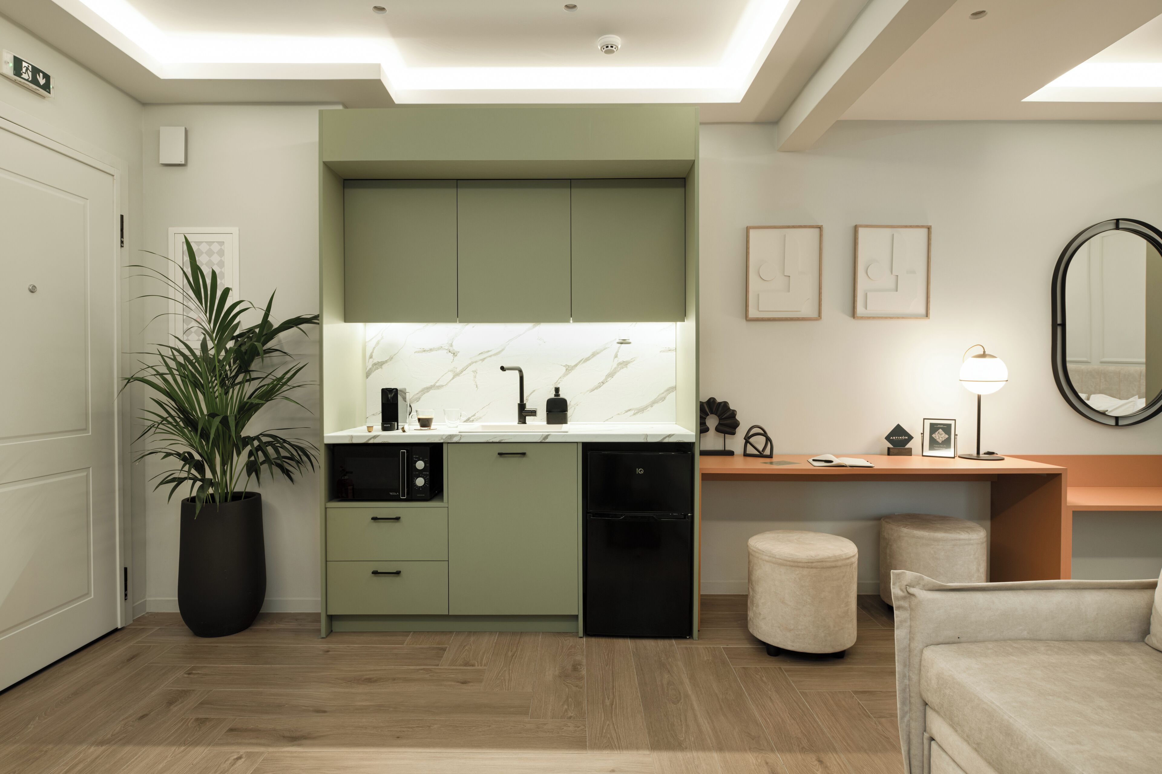 Een moderne keuken met groene kastjes, een marmeren spatwand en een zwarte vaatwasser. Er staat een plant in een pot, een klein rond tafeltje en een spiegel aan de muur.