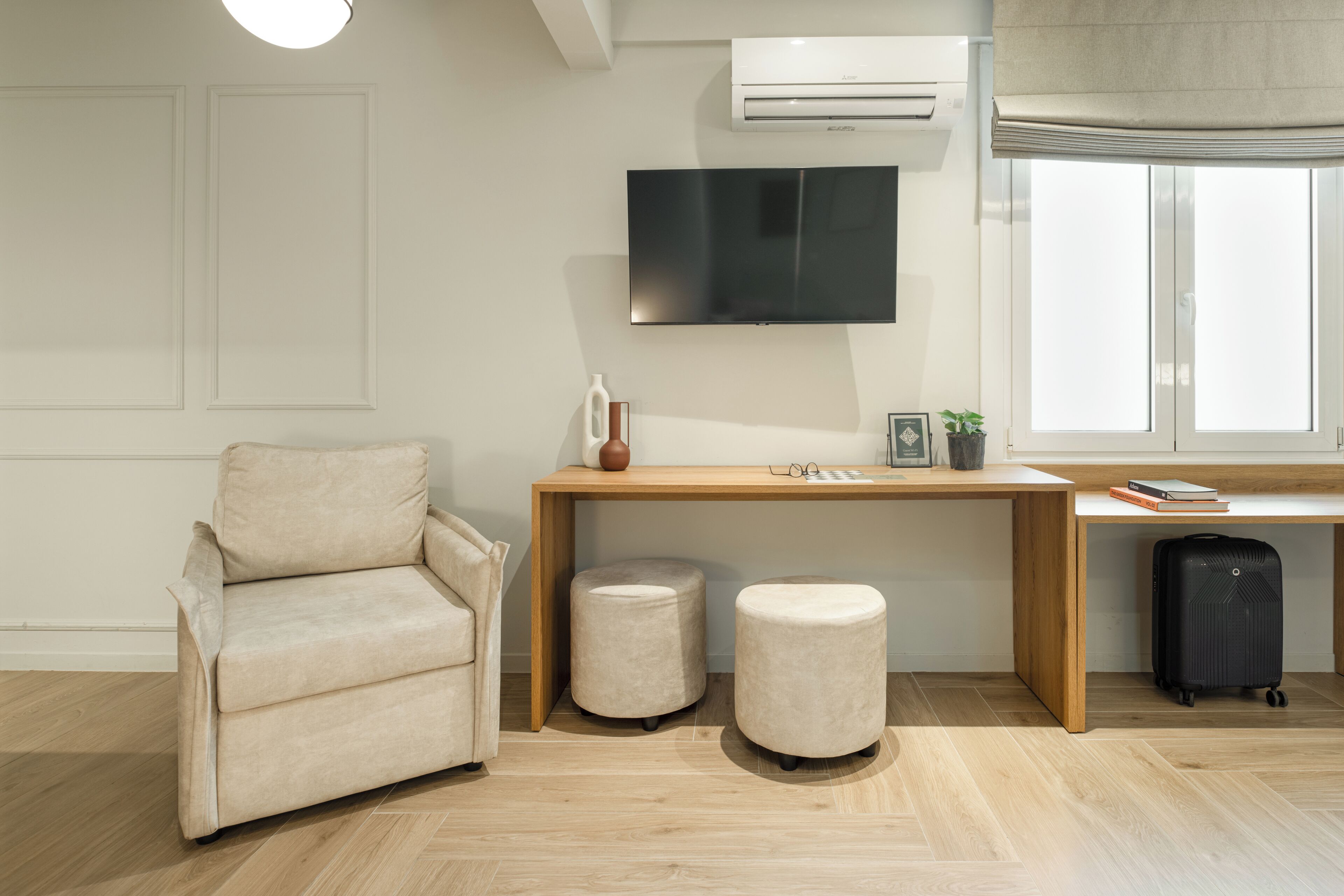 A modern living room with a sofa, a wooden desk, two ottomans, a television, and a suitcase.
