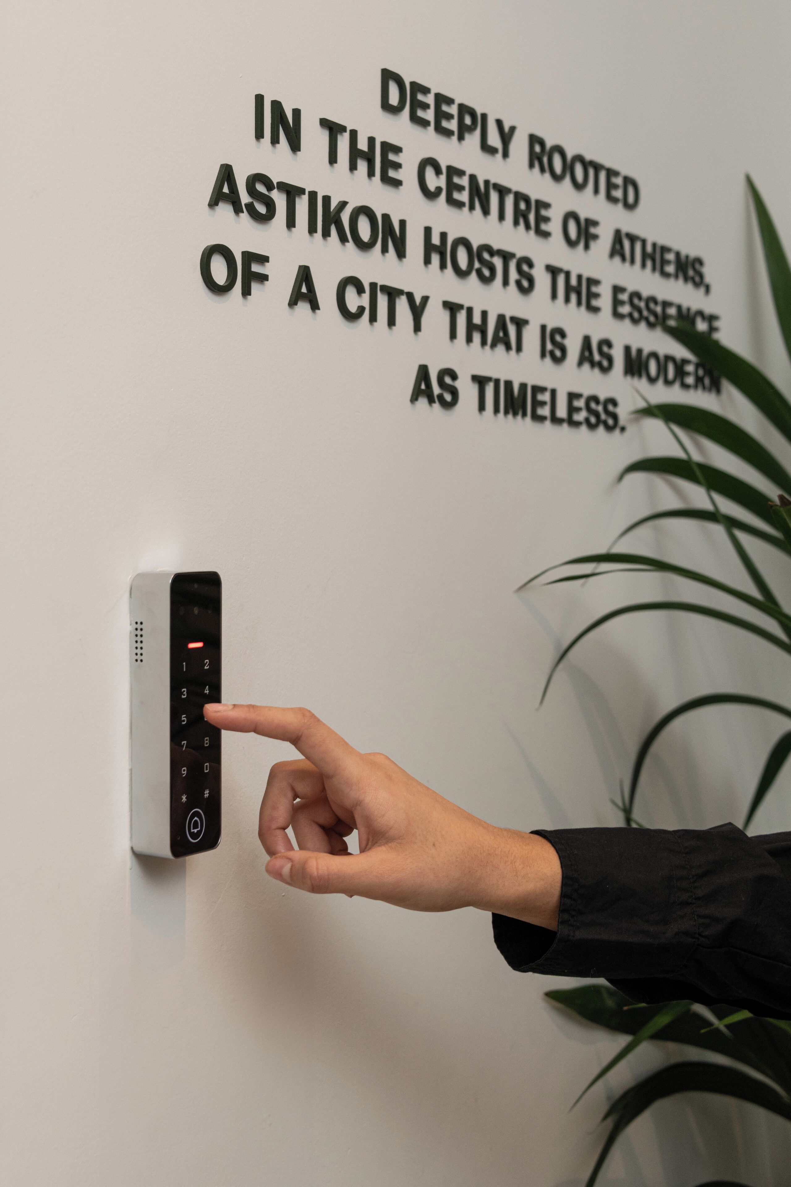A hand pressing a key on a wall-mounted device, with a sign stating "Deeply rooted in the centre of Athens, Astikon hosts the essence of a city that is as modern as timeless."