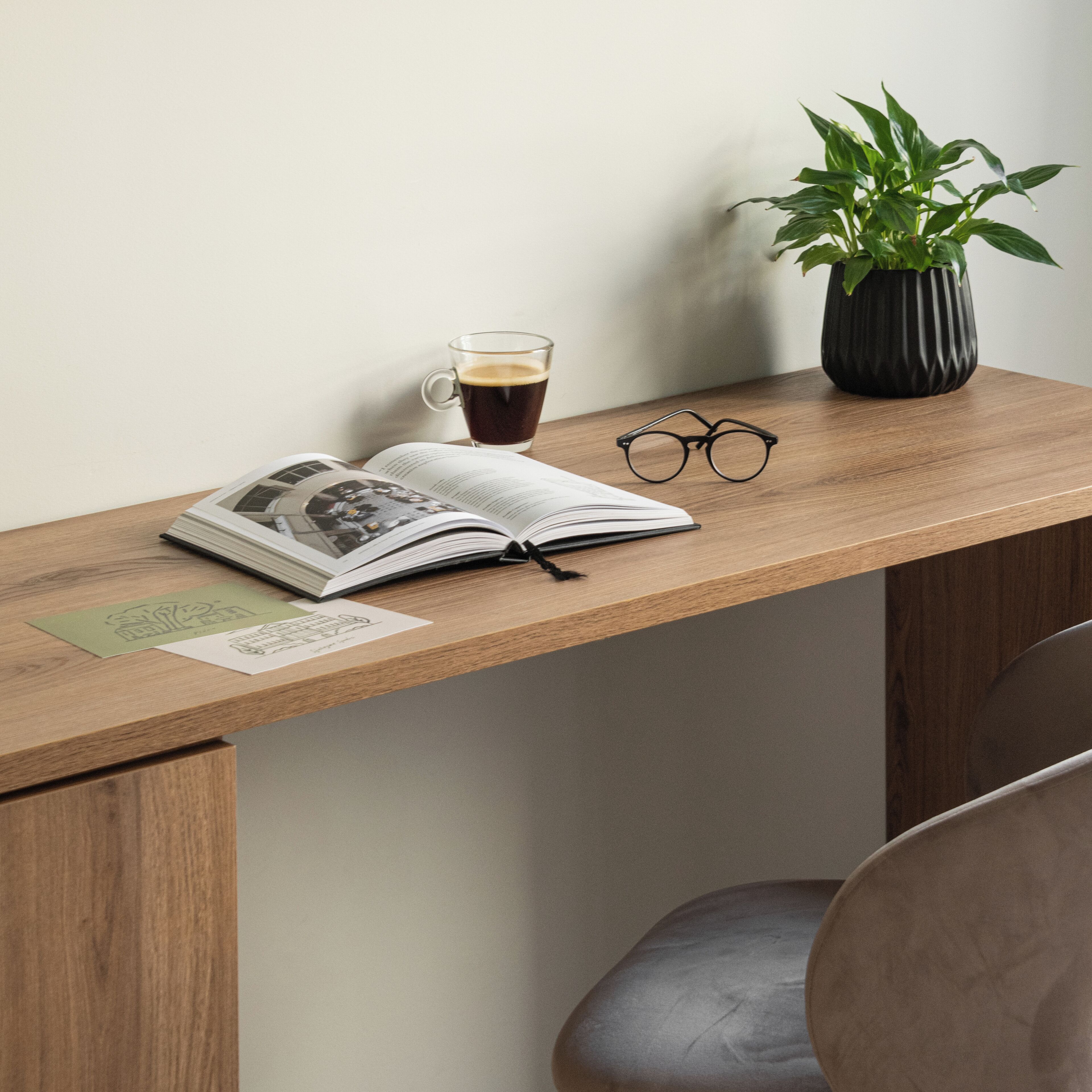 Un bureau en bois sur lequel se trouvent un livre ouvert, une tasse de café, une paire de lunettes et une plante en pot.