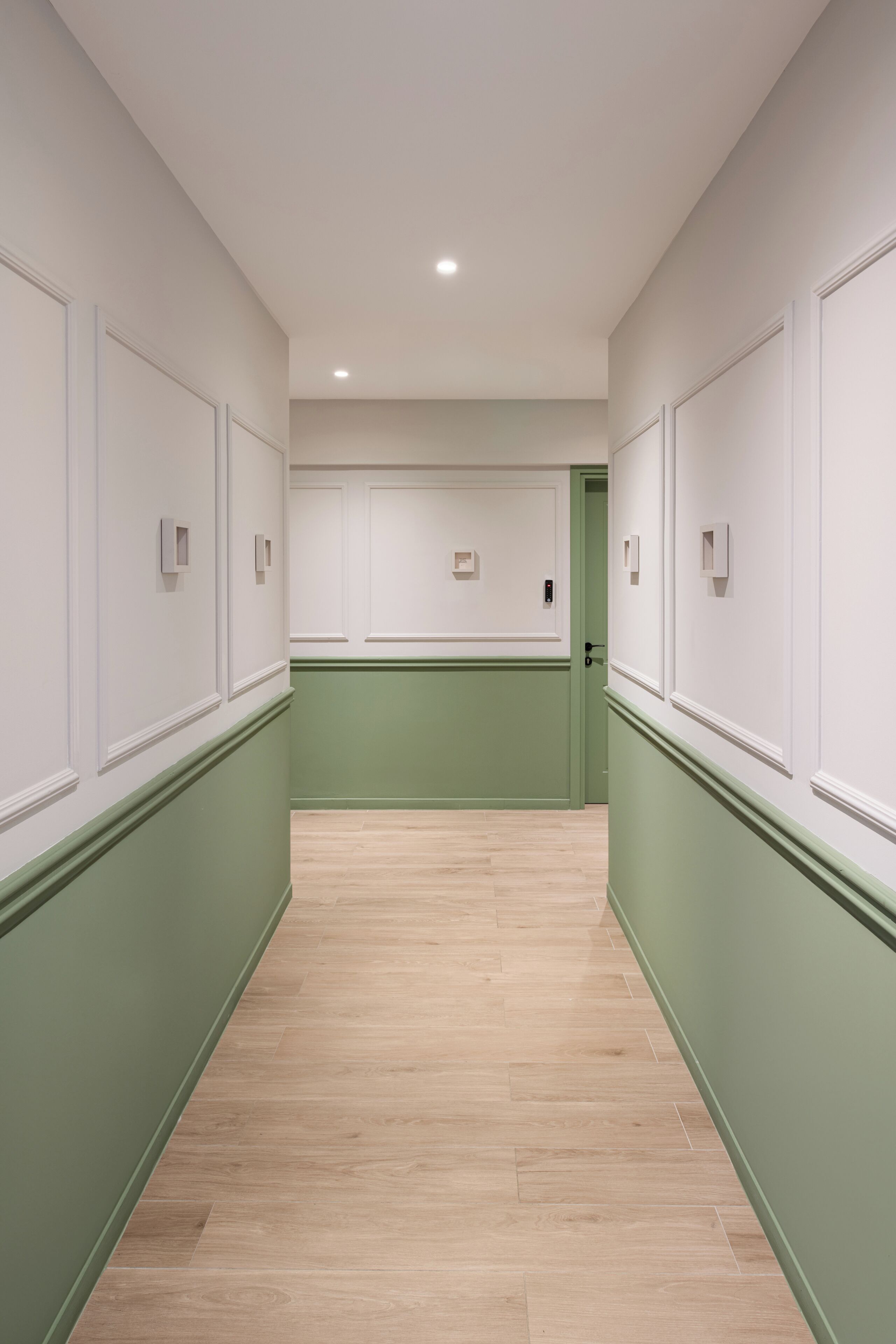 Un couloir aux murs verts, aux lambris blancs et au plancher en bois.