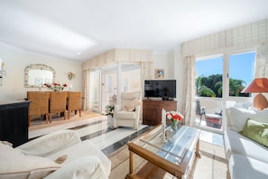 A spacious living room with a sofa, a coffee table, a TV, and a dining area with wicker furniture. - E04 Jardines 1 - Apartment (Sitio de Calahonda)