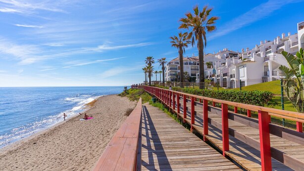 A wooden boardwalk along a beach with palm trees and white buildings in the background. - E04 Jardines 1 - Apartment (Sitio de Calahonda)