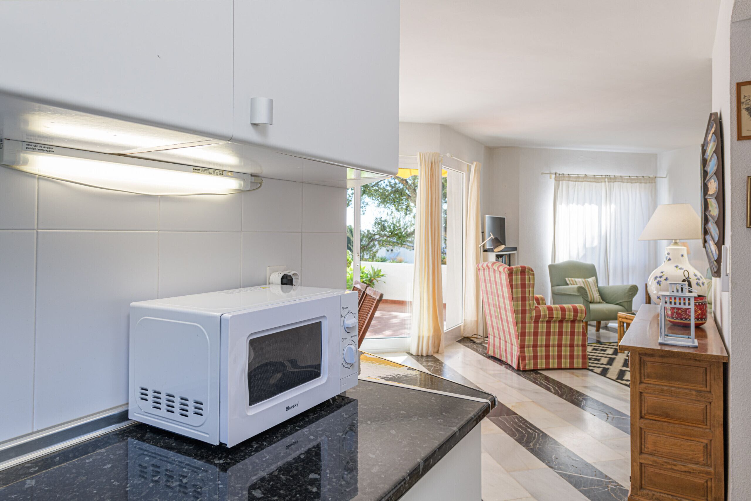 A kitchen with a microwave, a dining area with a chair, a lamp, and a view of the outdoors.
