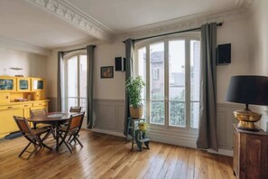 Ein Zimmer mit Holzboden, einem Esstisch mit Stühlen, einem gelben Schrank, einer Topfpflanze und einem Fenster mit Vorhängen.