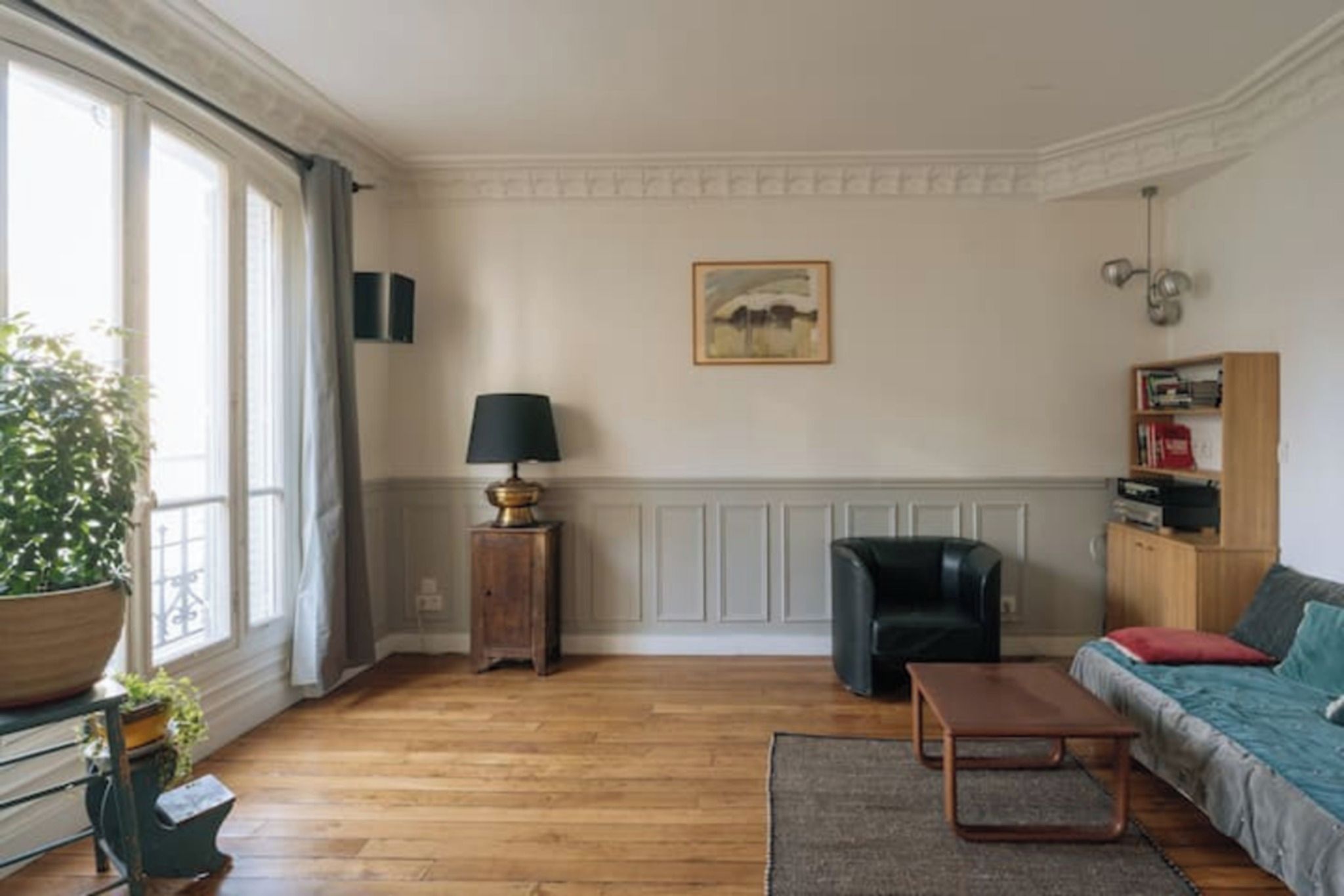 A living room with a wooden floor, a sofa, a coffee table, a lamp, a chair, a bookshelf, and a painting on the wall.