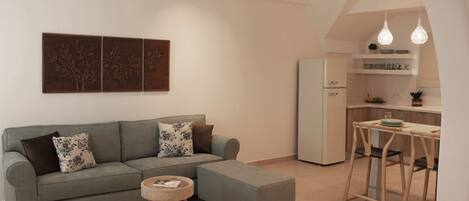 A modern living room with a grey sofa, ottoman, and round coffee table, a kitchen area with a white fridge and bar stools, and a dining table with hanging pendant lights.