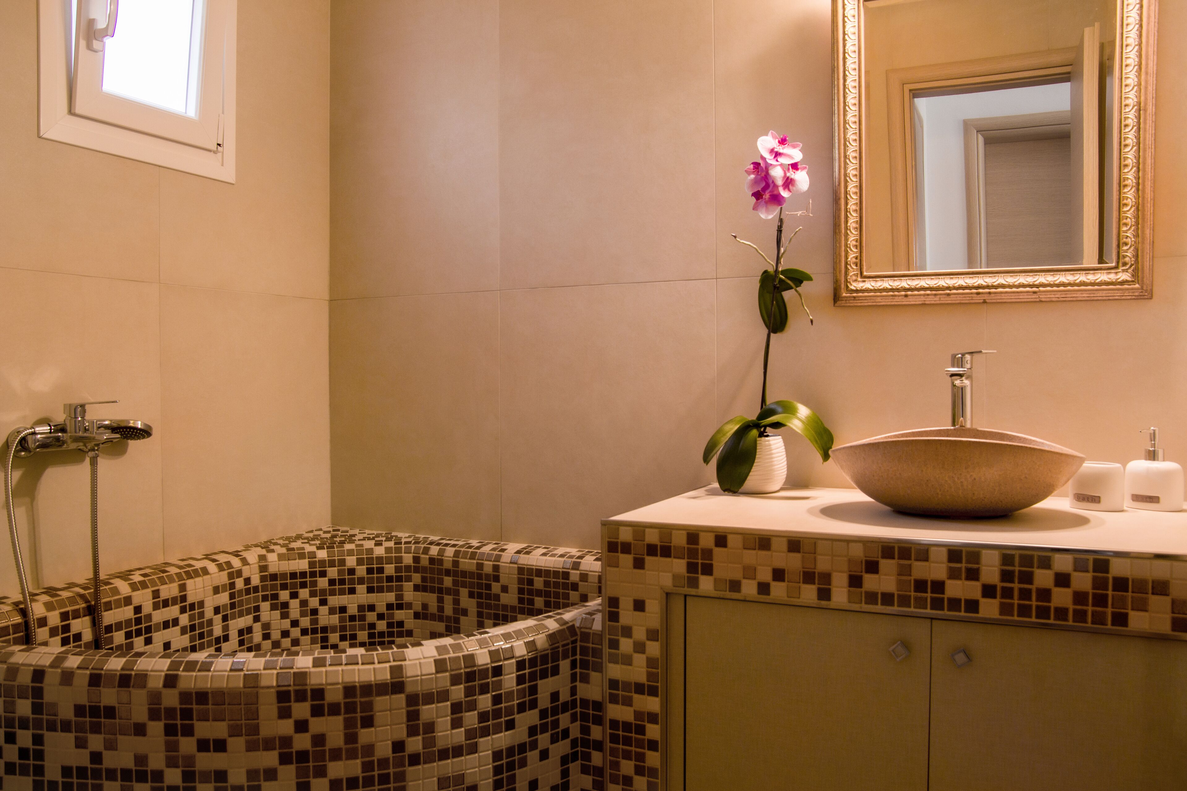 Un baño con bañera de mosaico, un lavabo, un espejo y una orquídea en maceta.