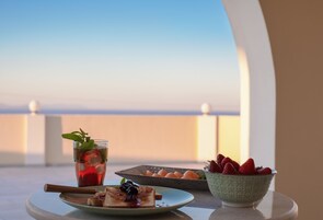 Una mesa con un vaso de té helado, un plato con tostadas y mermelada, un bol de fresas y un plato de pasteles en un balcón con vistas.