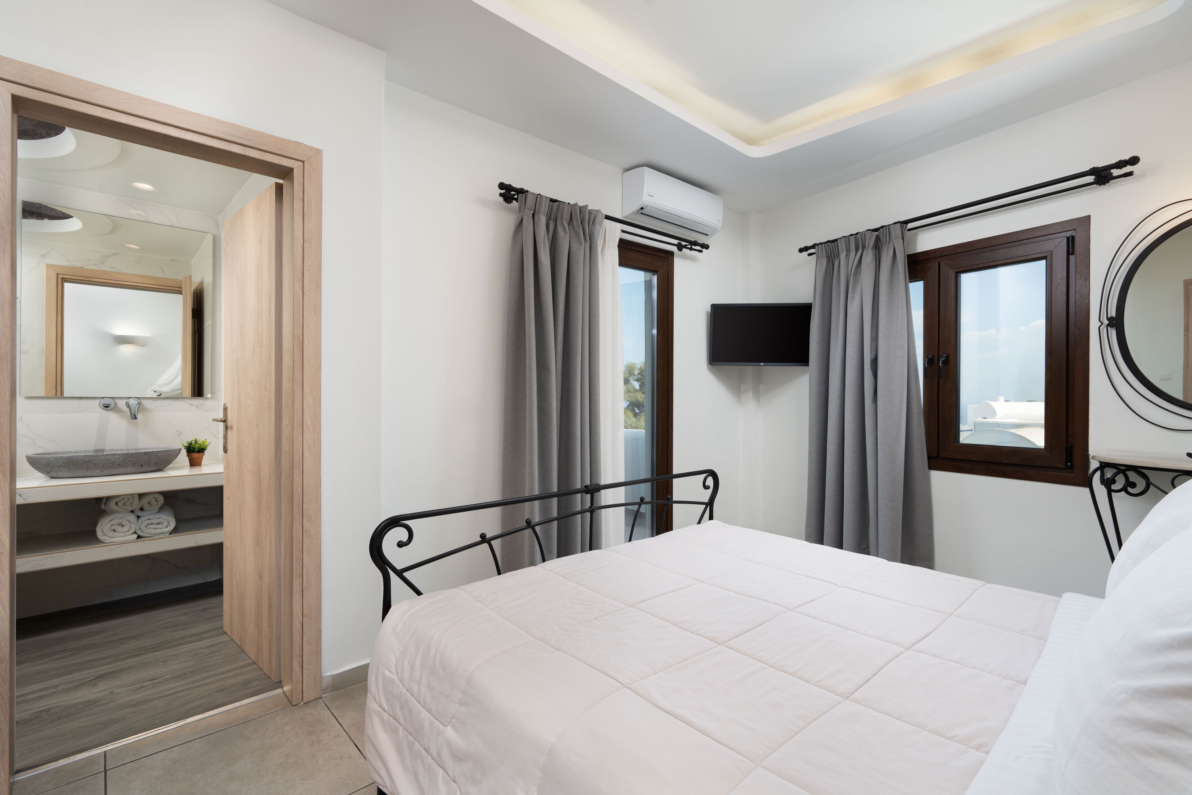Un dormitorio con cama, mesita de noche, televisor, aire acondicionado, ventana con cortinas y baño con lavabo y perchero para toallas.
