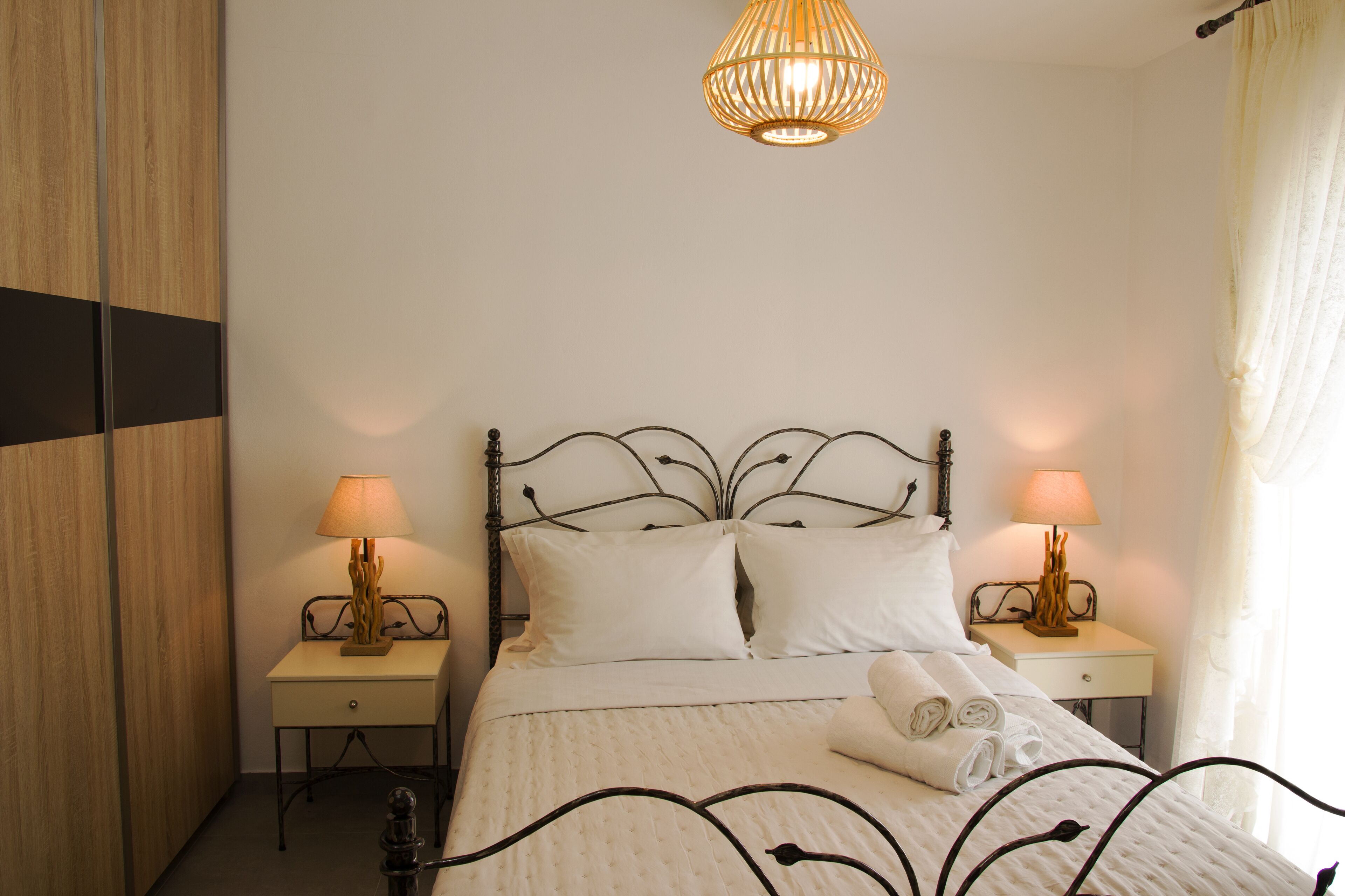 A neatly made bed with white linens and two bedside lamps. There is a wooden wardrobe to the left and a window with curtains to the right.