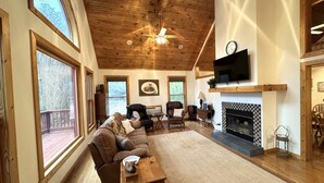 Un salon spacieux doté d’un grand plafond en bois, d’une cheminée, d’un téléviseur fixé au-dessus, et d’un coin salon confortable avec un canapé et des fauteuils.