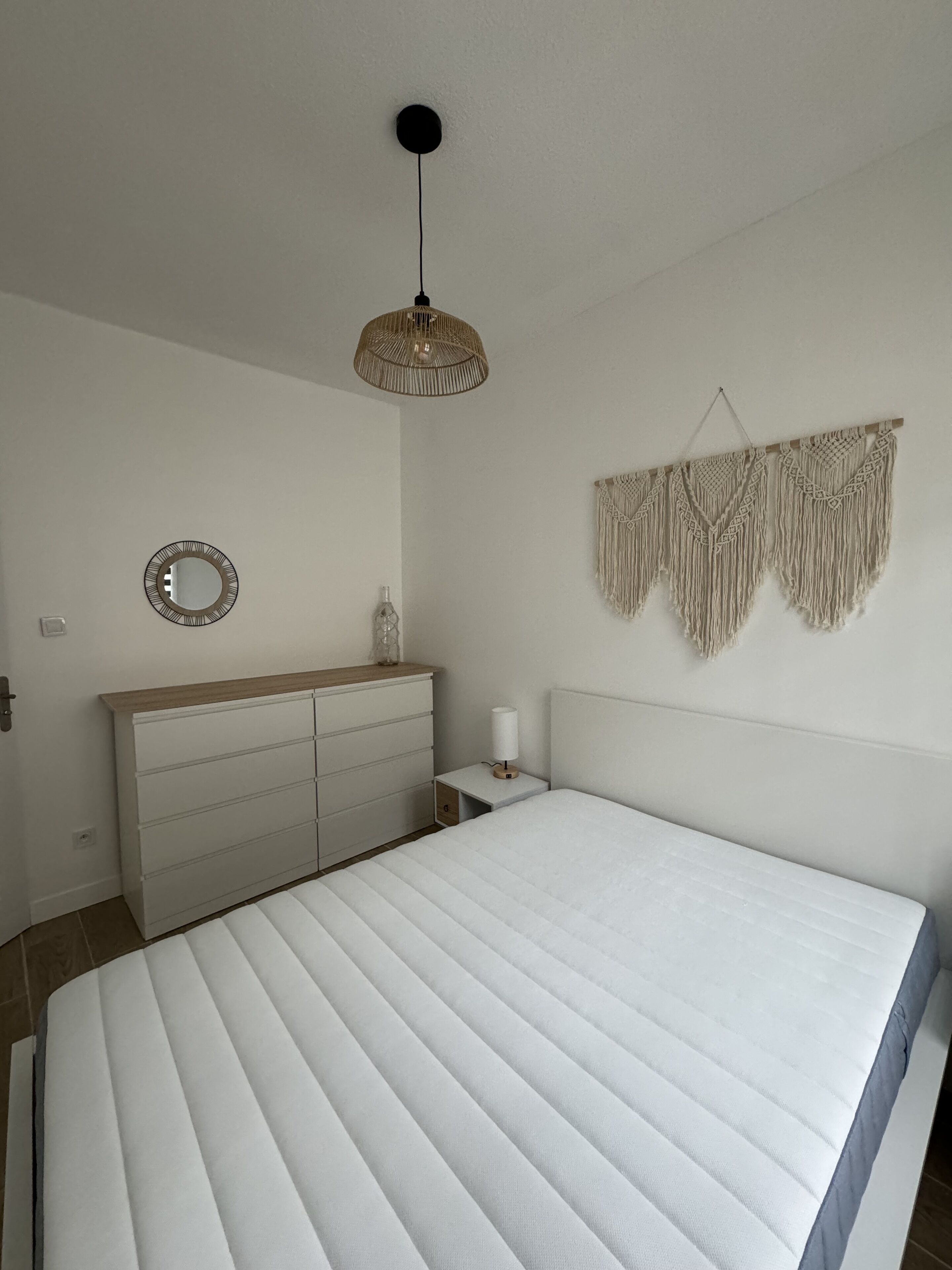 Ein Schlafzimmer mit einem großen Bett, einem Nachttisch mit Lampe, einem Schminktisch und einer Wanddekoration aus Makramee, sowie einer Deckenleuchte.