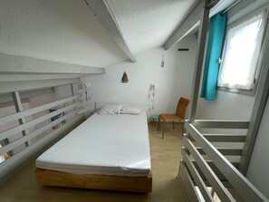 Een kleine kamer met een enkel bed, een houten hoofdbord, een stoel, een tafeltje en een raam met gordijnen.