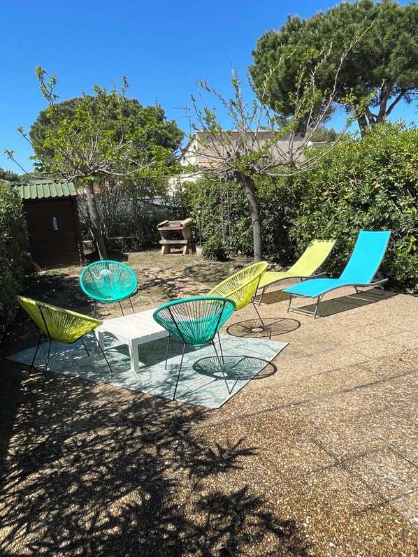 Een terras met tafel en stoelen, omringd door bomen en een klein gebouw.