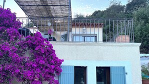 Een gebouw met twee verdiepingen, een balkon, een pergola en een grote struik paarse bloemen.