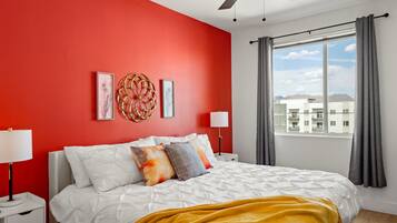 A bedroom with a red wall, a bed with white bedding, a yellow throw blanket, and a window with curtains.
