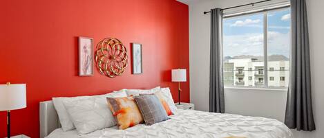 A bedroom with a red wall, a bed with white bedding, a yellow throw blanket, and a window with curtains.