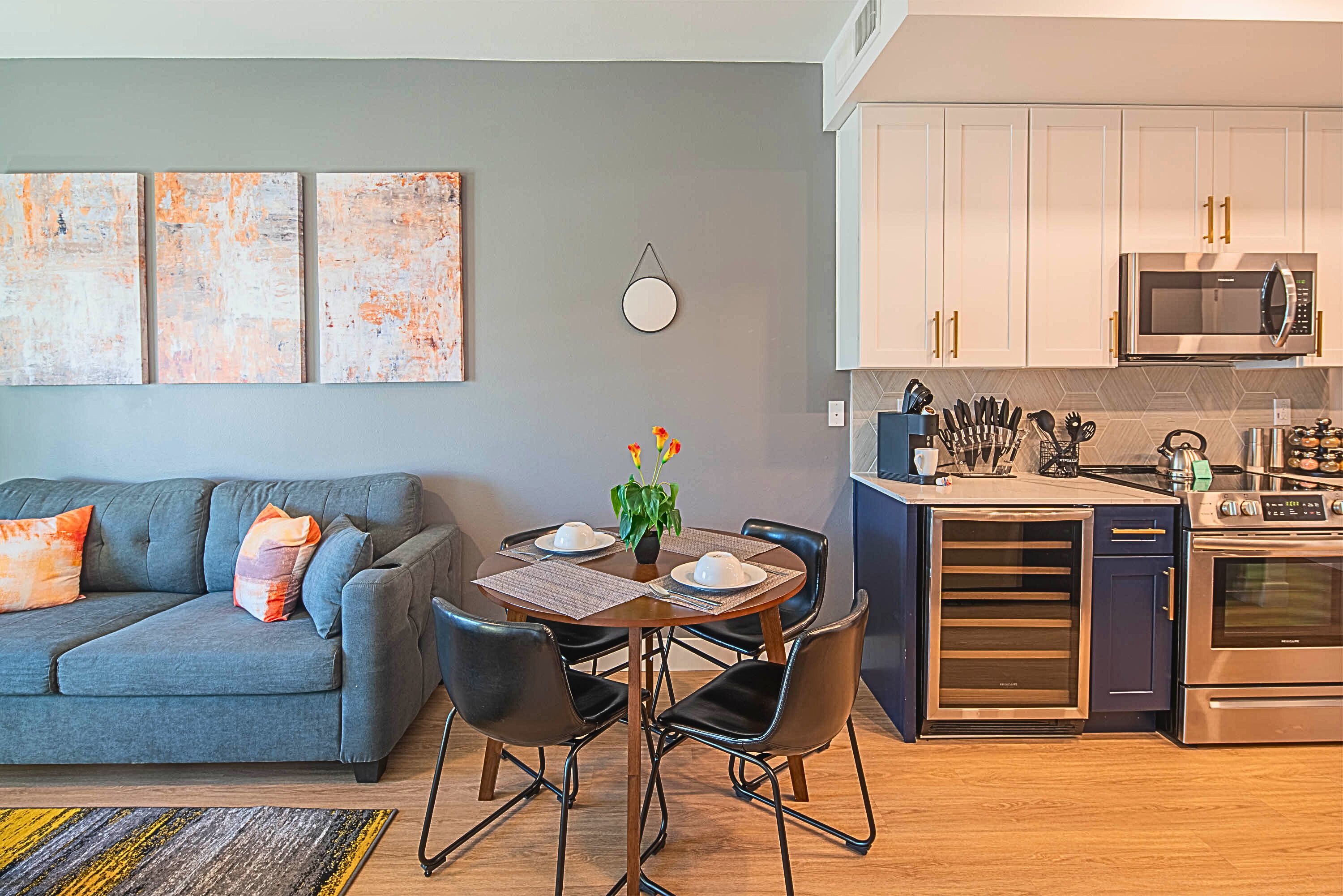 Una cocina moderna con electrodomésticos de acero inoxidable, una zona de comedor con mesa y sillas, y un sofá gris con cojines naranjas.
