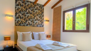 Un lit bien fait, avec du linge de lit blanc et une tête de lit beige, un mur orné d’un motif floral et une fenêtre donnant sur un paysage verdoyant.