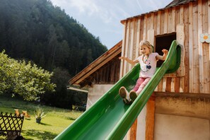 Un niño baja por un tobogán verde unido a una casita de juegos de madera con un adorno de flores.