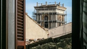 Blick durch ein Fenster mit einem Holzklappladen, der ein Baugerüst und eine untere Steinmauer zeigt.