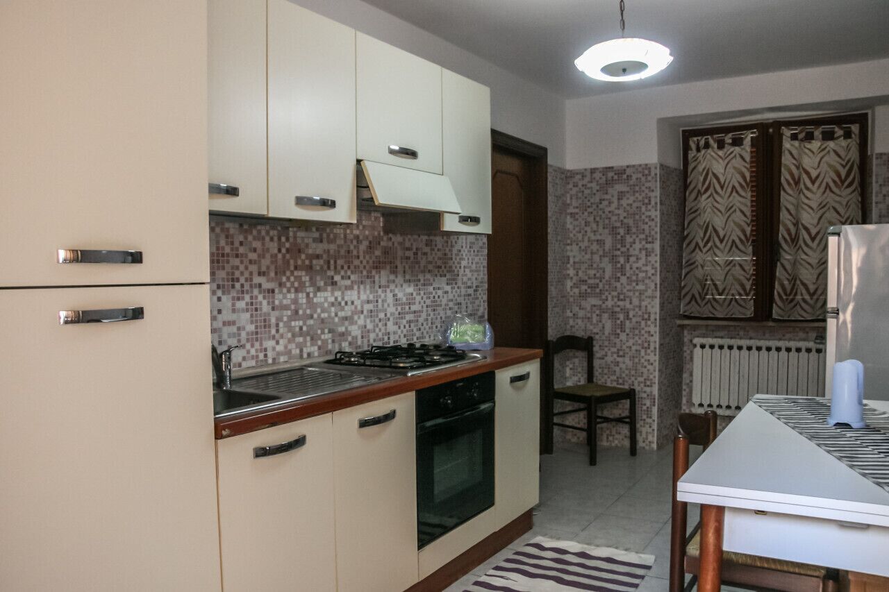 A compact kitchen with a gas stove, oven, and white cabinets. There is a dining table with chairs and a patterned rug.
