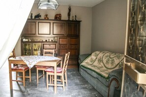 Un coin repas avec une table et des chaises en bois, un meuble contenant des objets décoratifs et un canapé vert agrémenté d’un plaid à motifs.