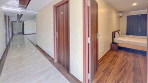 A hotel corridor with wooden doors, a bed with a blue curtain, and a tiled floor. - The Prime Hotel HBR (Bengaluru)