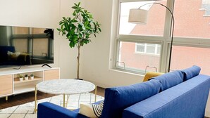 Una sala de estar moderna con un sofá azul, una mesa redonda blanca, un televisor sobre un mueble de madera y un amplio ventanal con vistas a edificios.