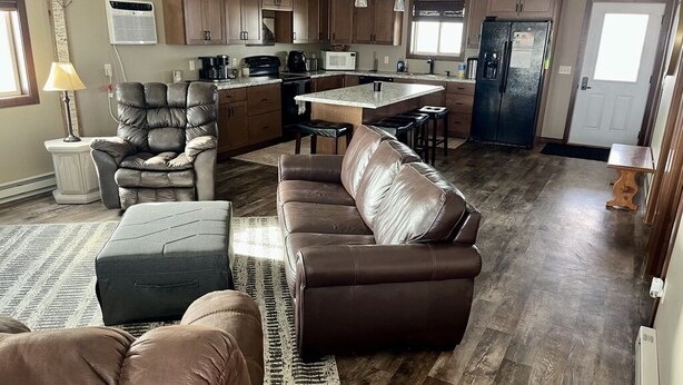 A spacious living room with a brown leather sofa, a matching ottoman, and a leather chair. The room features a large wooden floor, a kitchen area with wooden cabinets, and a dining area with a central island. - Quiet place on the prairie near Wessington, SD.  (Wessington)