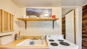 A compact kitchen with a white stove, a wooden countertop, a sink, and a shelf above holding dishes and a red cup. - 'Minnehaha' Allenspark Cabin Near Hiking! (Allenspark)