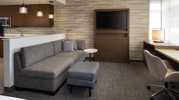 A modern hotel room with a grey sofa, a small ottoman, a desk with a chair, a flat-screen TV, and a kitchen area with a microwave.