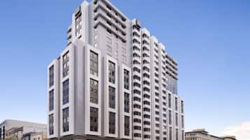 A modern high-rise building with a prominent corner design, multiple balconies, and a clear sky in the background.