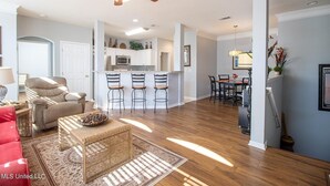 A spacious living room with a sofa, a coffee table, and a ceiling fan. There is a kitchen area with a microwave and a dining table with chairs.
