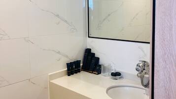 Un baño con pared de mármol blanco, un lavamanos blanco y un espejo. Hay artículos de aseo en el lavamanos.