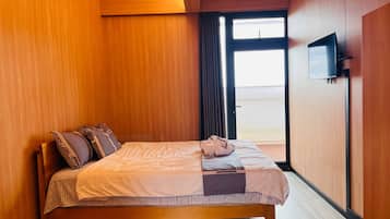 Una camera d'albergo con un letto in legno, una TV a schermo piatto fissata al muro e un'ampia finestra con tende.