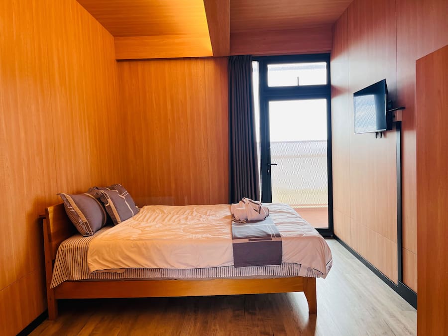 Quarto de hotel com cama de madeira, televisão de tela plana na parede e uma grande janela com cortinas.