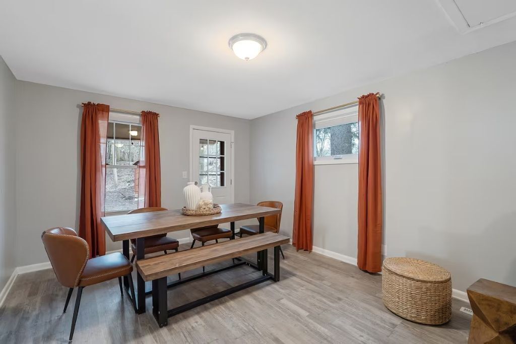 A dining area with a wooden table, benches, and chairs. There are two windows with orange curtains.