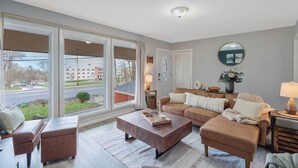 A spacious living room with a large window, a brown sofa set, a wooden coffee table, and a round mirror on the wall. - Hot Tub, Game Room, Near SU, sleeps 10 (Syracuse)
