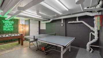 A ping pong table in a room with a neon sign, a foosball table, and a sign indicating a game room.