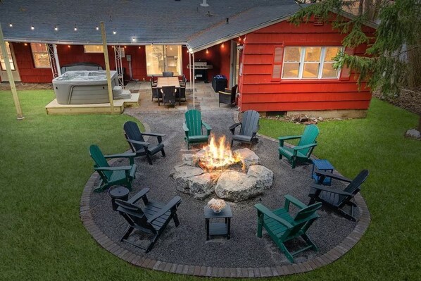 A circular seating area with fire pit and Adirondack chairs, a hot tub, and a red building with a covered patio. - Hot Tub, Game Room, Near SU, sleeps 10 (Syracuse)