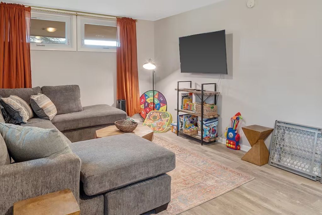 Ein Wohnzimmer mit grauem Sofa, Hocker und einem an der Wand montierten Fernseher. Ein Bücherregal beherbergt Spielzeug und ein kleiner Tisch mit Lampe steht daneben.