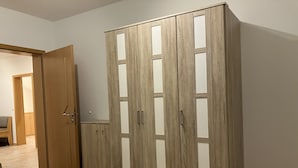 Un armario de madera con varios compartimentos y un cajón pequeño, situado en un pasillo con suelo de baldosas y una puerta que da a otra habitación.