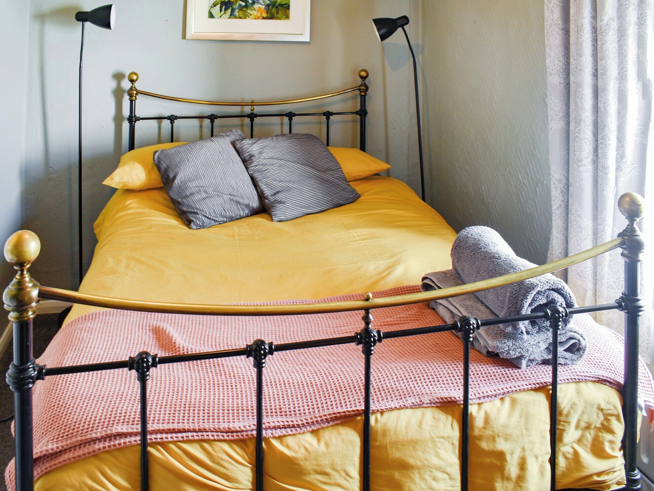 A bed with a yellow bedspread, a pink knitted blanket, and a grey towel.