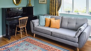 A living room with a grey sofa, a wooden chair, a black piano, a patterned rug, and a window with curtains. - 4 bedroom accommodation in Saxmundham (Saxmundham)