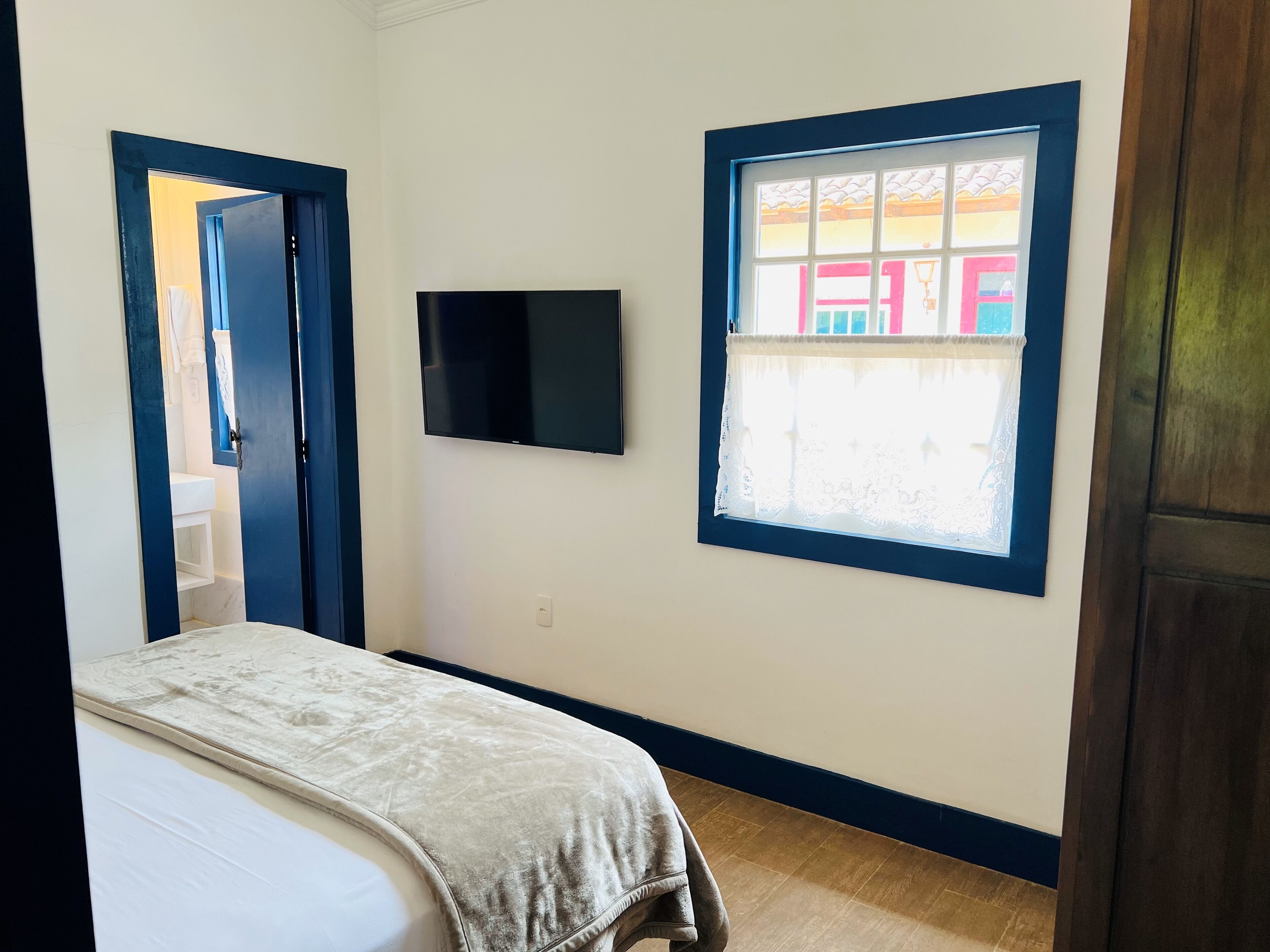 A bedroom with a bed, a TV mounted on the wall, and a window with a view of colorful buildings.